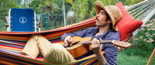 Ein Mann mit Strohhut entspannt in einer bunten Hängematte, spielt Gitarre. Im Hintergrund ist ein Garten mit Blumen sichtbar.