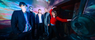 Fünf Personen posieren vor einer farbenfrohen, künstlerischen Wand. Eine Person trägt Rot und hebt den Arm.