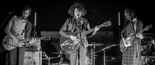 Drei Musiker spielen auf einer Bühne: Zwei spielen elektrische Gitarren, eine Person singt ins Mikrofon; es ist schwarz-weiß.