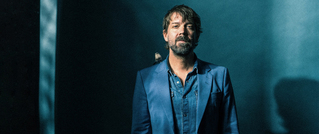 Ein Mann in einem blauen Anzug steht vor einem dunkelblauen Hintergrund. Auf seiner Schulter sitzt ein kleiner Vogel.