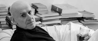 Ein älterer Mann sitzt auf einem Sofa, hält ein Getränk. Im Hintergrund sind gestapelte Bücher zu sehen.