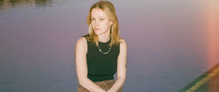 Eine Person mit hellem Haar sitzt am Wasser, trägt ein schwarzes Oberteil und eine bunte Halskette. Der Hintergrund ist verschwommen.