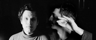 Zwei Männer in Schwarz-Weiß-Porträt; einer schaut nach vorn, der andere zur Seite mit erhobener Hand.