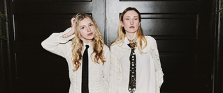 Zwei Personen in weißen Hemden und Krawatten stehen vor einer dunklen Tür. Eine Person berührt ihr Haar, die andere blickt ernst.