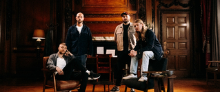 Vier Personen in einem eleganten holzgetäfelten Raum, sitzen und stehen lässig. Die Atmosphäre wirkt ruhig und stilvoll.