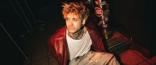 Ein Mann mit zerzaustem Haar trägt eine rote Jacke und sitzt auf dem Boden. Der Hintergrund zeigt einen mit Aufklebern bedeckten Automaten.