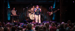 Eine Band spielt auf einer Bühne vor einem großen Publikum. Ein Sänger steht im Vordergrund, mit einer Band im Hintergrund.