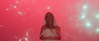 Frau in weißem Kleid mit Schleier vor rotem, funkensprühenden Hintergrund, sieht verträumt aus.