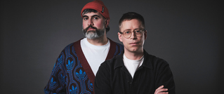 Zwei Personen vor dunklem Hintergrund. Eine trägt Brille und dunkle Kleidung, die andere Mütze und ein gemustertes Oberteil.