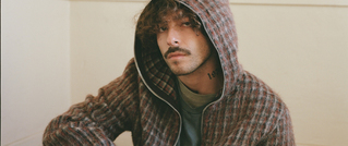 Junger Mann mit lockigem Haar und Schnurrbart sitzt entspannt. Er trägt einen karierten Hoodie und hat ein Tattoo am Hals.