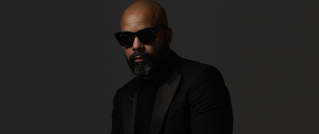 Eine Person mit Glatze und Bart trägt eine schwarze Sonnenbrille und einen schwarzen Anzug vor einem dunklen Hintergrund.