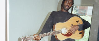 Person in dunklem Pullover hält eine Akustikgitarre mit der Aufschrift 'EXEX'. Hintergrund zeigt ein Fenster mit Bäumen draußen.