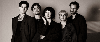 Eine Gruppe von fünf Personen in eleganter Kleidung posiert vor einem neutralen Hintergrund, alle schauen in die Kamera.