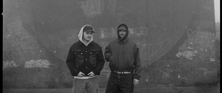 Zwei Personen stehen vor einer Betonwand, beide tragen Kapuzenpullis. Das Bild ist in Schwarzweiß gehalten.