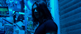 Eine Person mit langen Haaren steht in einem blauen, neonbeleuchteten Raum mit Postern an der Wand.