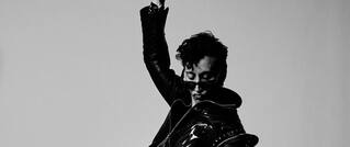 Person in schwarzer Lederjacke hebt eine Hand. Das Bild ist in Schwarz-Weiß gehalten mit einem minimalistischen Hintergrund.