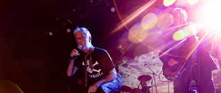 Zwei Musiker auf einer Bühne, einer singt ins Mikrofon, der andere spielt Bass. Bunte Lichtreflexe und ein Schlagzeug im Hintergrund.