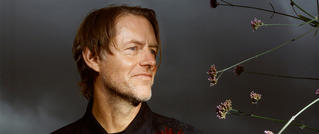 Ein Mann mit schulterlangem Haar schaut lächelnd zur Seite. Im Vordergrund sind dünne Äste mit kleinen Blüten sichtbar.