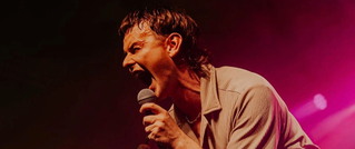 Ein Sänger mit nassen Haaren und beigem Hemd schreit leidenschaftlich ins Mikrofon vor einem pinken, beleuchteten Hintergrund.