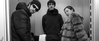 Drei Personen posieren in einem Aufzug. Alle tragen stilvolle Winterkleidung, darunter eine Pelzjacke und Sonnenbrillen. Schwarz-Weiß-Foto.
