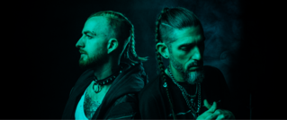 Zwei Männer mit geflochtenem Haar in einem grünen Licht. Beide blicken nach links, der Hintergrund ist dunkel und rauchig.