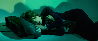 Person liegt in grünem Licht neben einem Keyboard und Verstärker, mit dem Kopf auf einem Turnschuh ruhend, in nachdenklicher Pose.