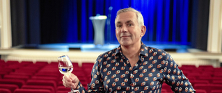 Eine Person sitzt in einem leeren Theater mit roten Sitzen, hält ein Weinglas und trägt ein gemustertes Hemd, Bühne im Hintergrund.