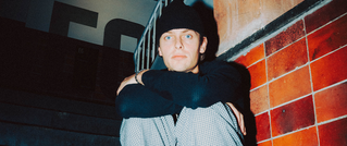Ein Mann sitzt in einem Treppenhaus, trägt eine schwarze Mütze und Pullover, und hat die Arme um die Knie geschlungen. Im Hintergrund sind rote Fliesen.