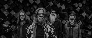 Vier Personen stehen vor einer Wand aus gestapelten Holzwürfeln. Drei tragen lange Haare, eine Person eine Sonnenbrille. Schwarzes Weiß.