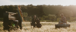 Drei Musiker spielen im Freien: einer am Klavier, einer mit Cello, der dritte mit Gitarre auf einem Teppich. Hintergrund: Bäume.