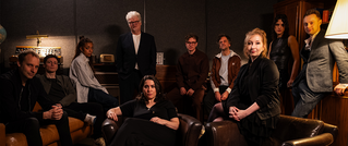 Zehn Personen sitzen und stehen in einem gemütlich beleuchteten Raum mit Sesseln, Regalen und einer Lampe. Alle schauen in die Kamera.