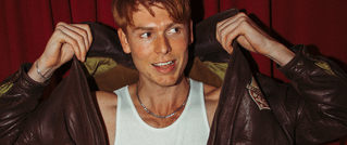 Person mit rostrotem Haar öffnet eine Lederjacke, darunter ein weißes Tanktop. Roter Vorhang im Hintergrund.