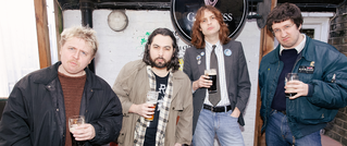 Vier Männer stehen in lässiger Kleidung draußen und halten Biergläser in den Händen. Im Hintergrund ist ein Guinness-Schild.