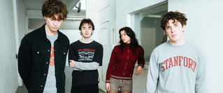 Vier Personen stehen in einem weißen Flur. Drei schauen ernst, eine Frau trägt Rot. Einer trägt ein graues 'Stanford'-Sweatshirt.