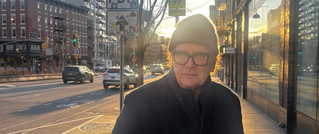 Ein Mann mit Brille und Mütze steht auf einer sonnigen Stadtstraße, links sind Autos und Gebäude zu sehen.