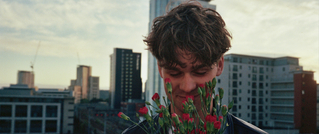 Ein Mann riecht an einem Strauß roter Nelken auf einem Dach, im Hintergrund eine Skyline bei Sonnenuntergang.