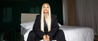 Person mit langen blonden Haaren sitzt in schwarzem Anzug auf einem Bett in einem dunklen Raum.