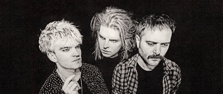 Drei Männer in Schwarz-Weiß, mit unterschiedlicher Frisur und Gesichtsausdruck, schauen in verschiedene Richtungen.