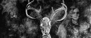 Person hält einen Hirschschädel mit großen Geweihen, umgeben von Rauch. Im Hintergrund sind zwei unscharfe Gestalten zu sehen.