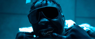 Person mit großer Sonnenbrille und Grills auf den Zähnen lächelt im blauen Licht. Sie trägt eine dicke Jacke.