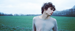 Eine Person mit lockigem Haar steht in einem grauen Shirt auf einem Feld. Der Himmel ist bewölkt, und im Hintergrund sind Bäume.