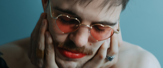 Person mit roten Lippen und rosafarbenen Sonnenbrillen hält ihr Gesicht in den Händen. Hintergrund ist blau.