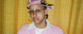 Person mit Brille trägt eine rosa Kappe und ein rosafarbenes Oberteil. Gelber Vorhang im Hintergrund.