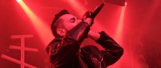 Ein Sänger in einer Lederjacke performt auf einer Bühne im roten Licht, hält ein Mikrofon nah an den Mund.