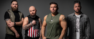 Vier Männer in lässiger Kleidung posieren vor grauem Hintergrund. Einer trägt ein USA-Flagge-T-Shirt, ein anderer ein grünes Shirt.