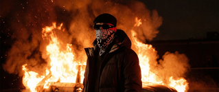 Eine vermummte Person steht vor einem brennenden Auto in der Nacht. Flammen lodern hell und erleuchten die Szene.