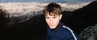 Ein junger Mann in blauer Jacke steht draußen vor einer Berglandschaft im Hintergrund.