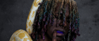 Person mit bunten Dreadlocks hält einen gelben Python. Trägt violetten Zahnschutz und posiert vor grauem Hintergrund.