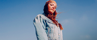 Frau mit roten Haaren und Stirnband in Jeansjacke, schaut nachdenklich in die Ferne vor klarem blauem Himmel.