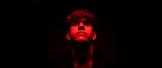 Mann mit Locken schaut in rotem Licht geradeaus. Schwarzer Hintergrund und Schatten betonen Gesichtszüge.